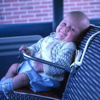 A baby sits in an unsafe, old metal car seat positioned in the front smiling toward the camera.
